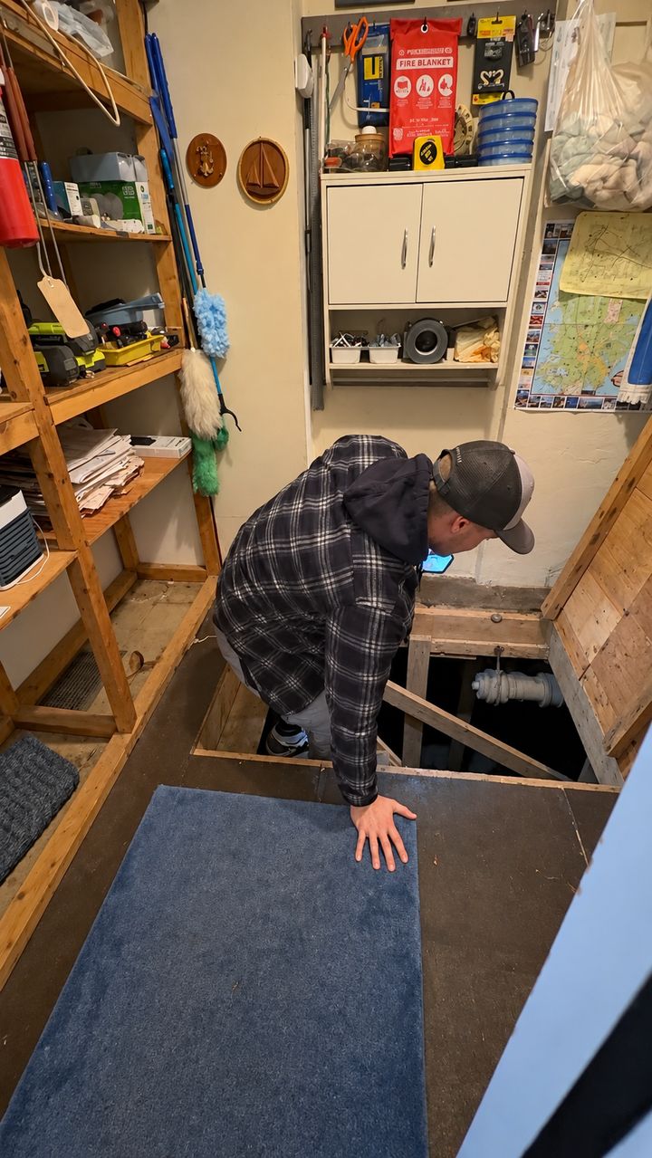 Greg Valentgas looking through a floor hatch in a utility room to check below the house